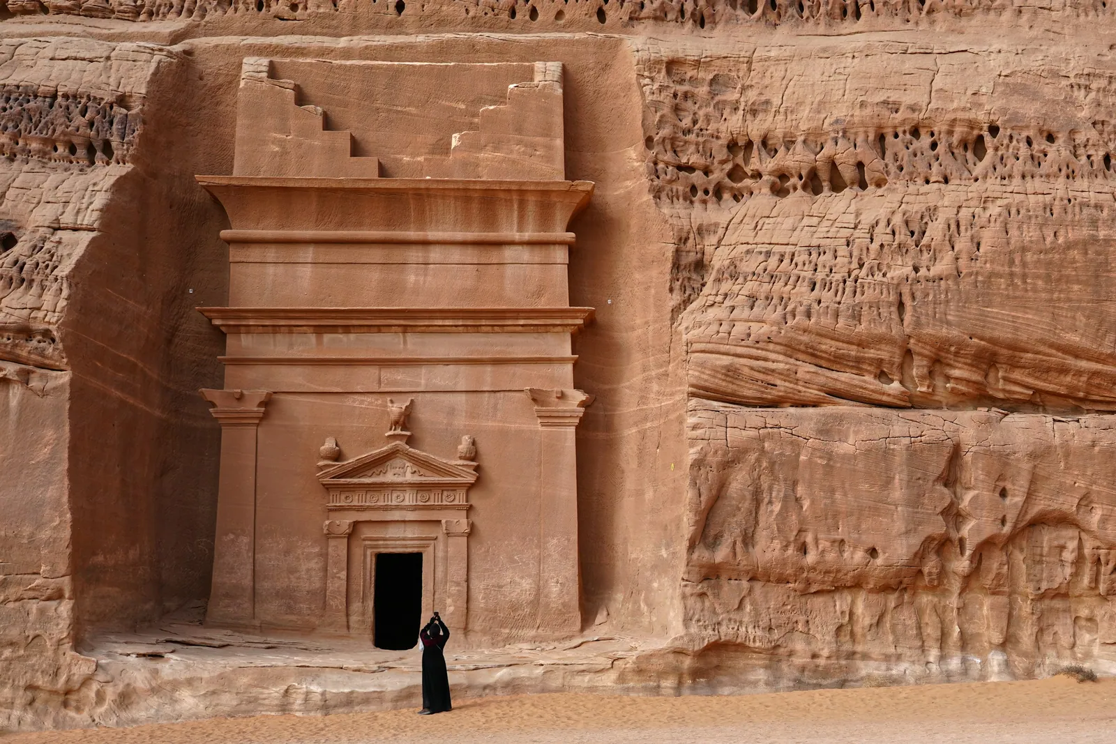 A Nabataean tomb facade carved into a sandstone cliff at Hegra in AlUla, with a lone visitor standing at its doorway