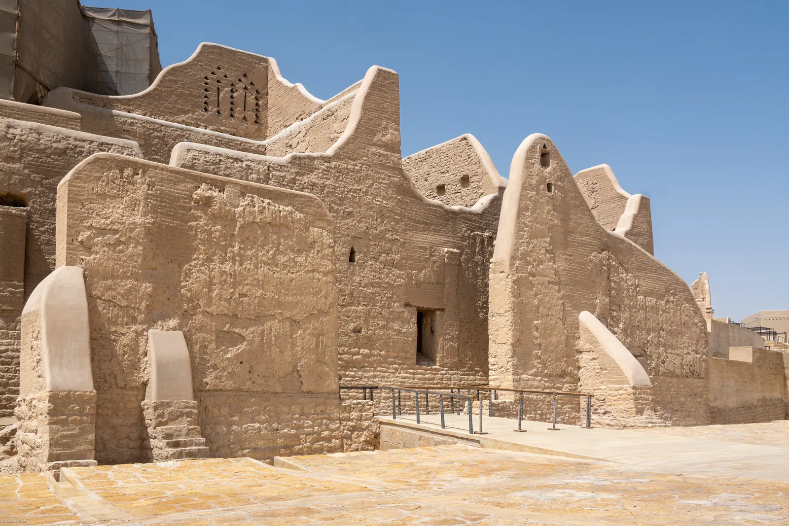 The restored mud-brick walls and triangular crenellations of At-Turaif in Diriyah under a clear blue sky, with a walkway between the buildings