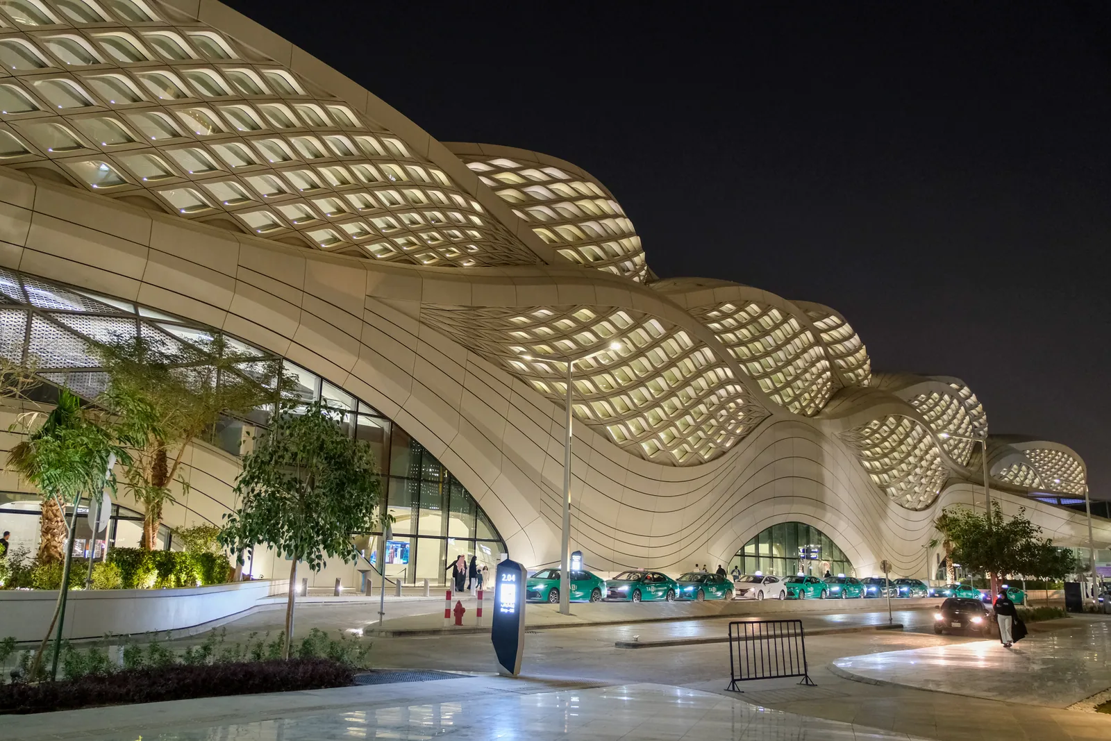 The KAFD metro station in Riyadh at night, its flowing latticed roof by Zaha Hadid Architects lit from within, with taxis waiting outside