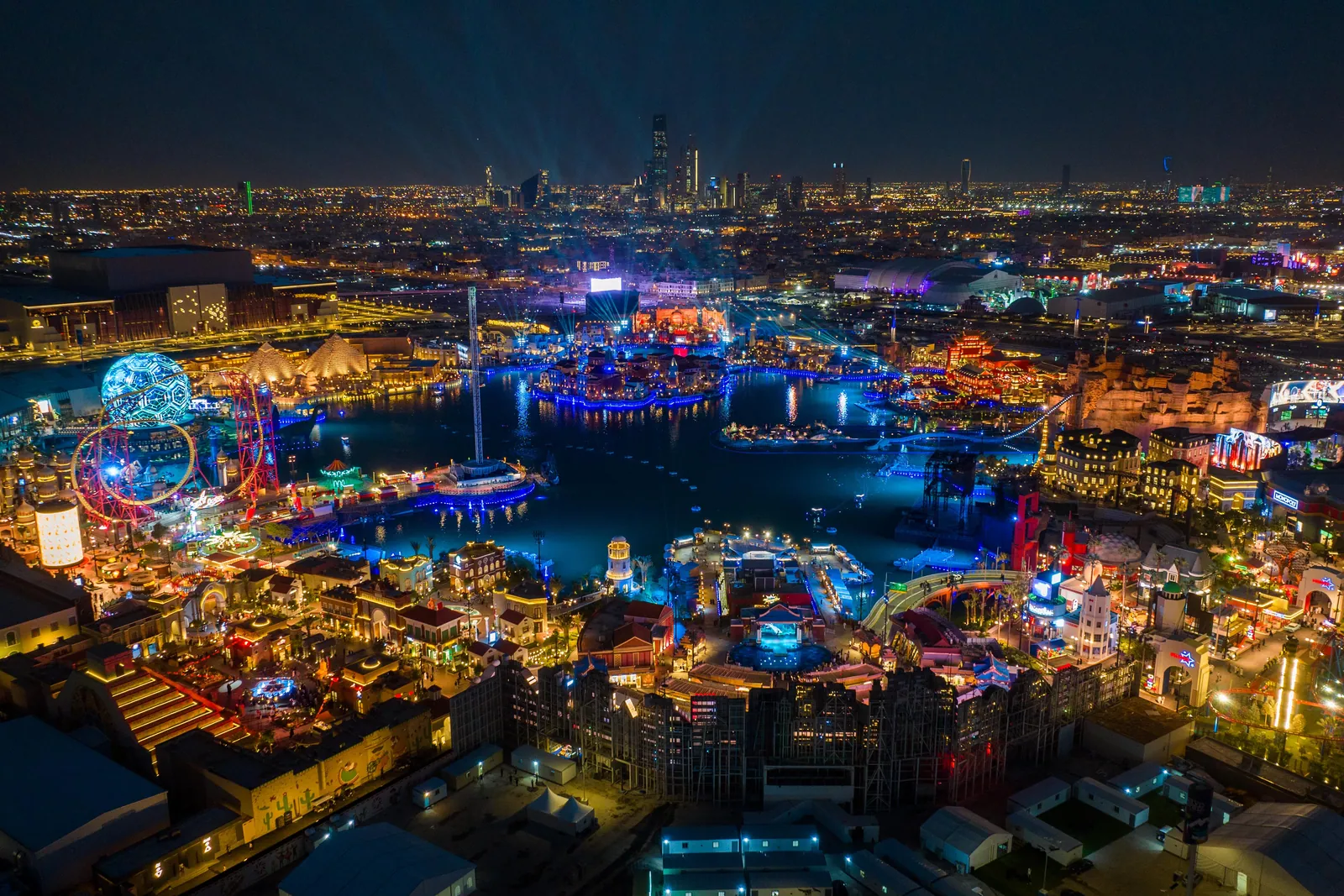 Aerial view of Boulevard World at night during Riyadh Season, its lagoon and country-themed districts lit up, with the Riyadh skyline behind
