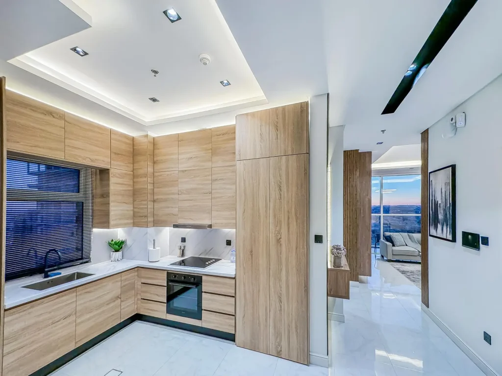 Modern U-shaped kitchen in light wood and marble finishes, opening to hallway views of the high-floor furnished apartment with balcony.