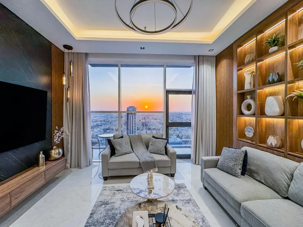 Sunset framed through panoramic windows, flanked by neutral sofas and accent lighting in a high-floor furnished apartment with balcony.